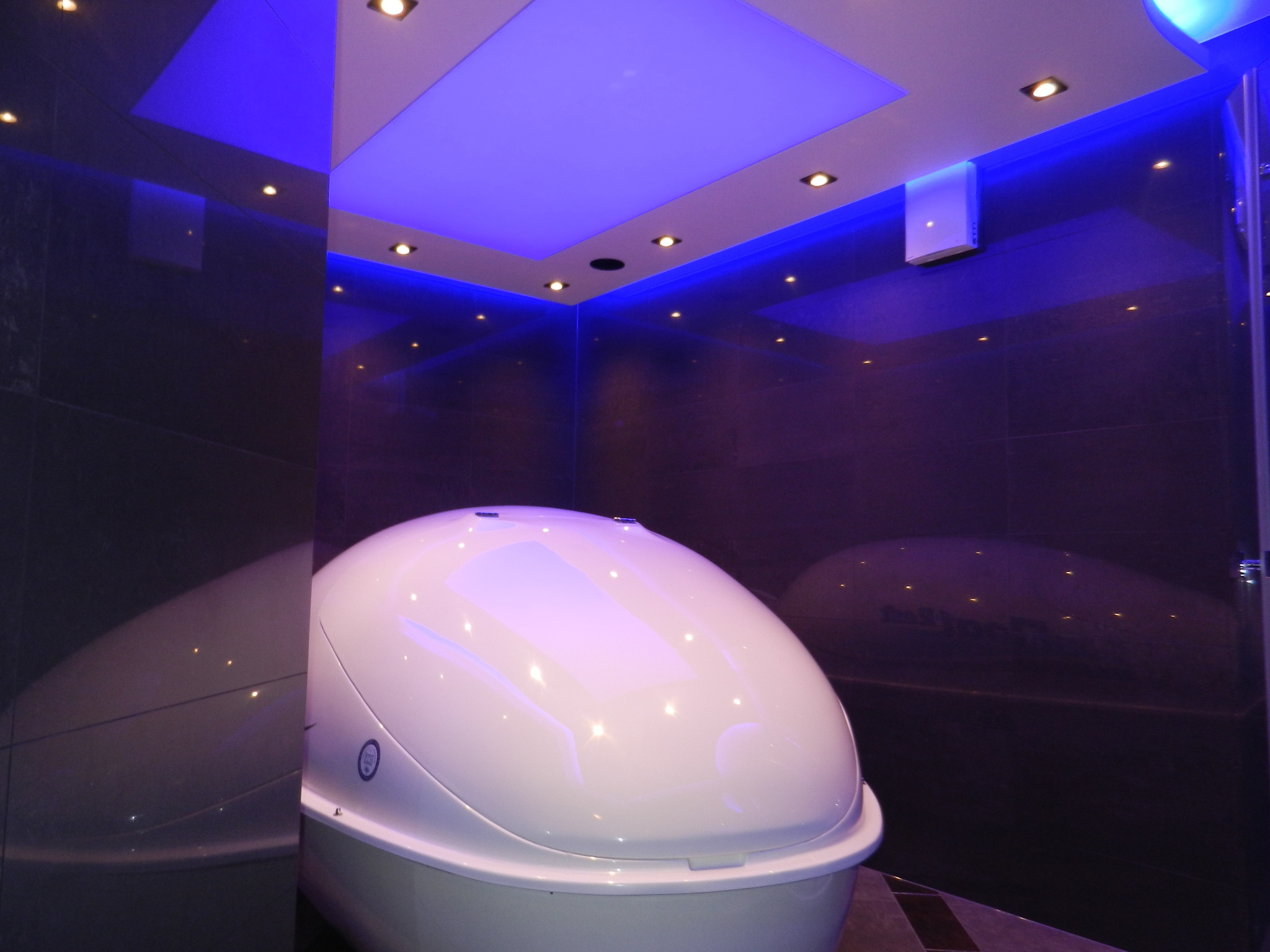 Nowoczesna kapsuła sensoryczna w pomieszczeniu z nastrojowym, niebieskim oświetleniem punktowym i kwadratowym panelem sufitowym.