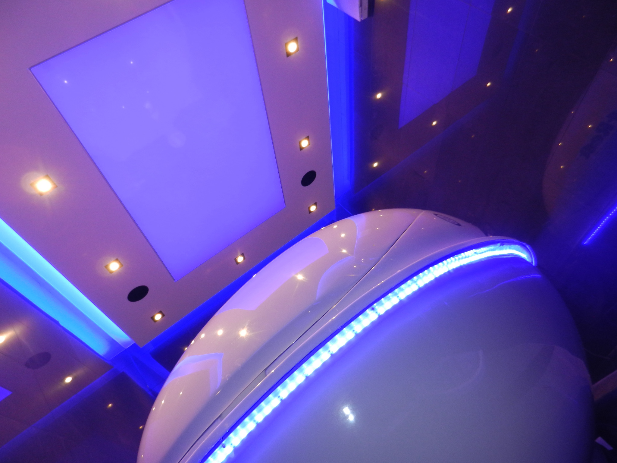 Wnętrze kabiny floatingowej z podświetleniem LED, widok z dołu na białą kapsułę i sufit z lampami punktowymi i podświetleniem w kolorze niebieskim.