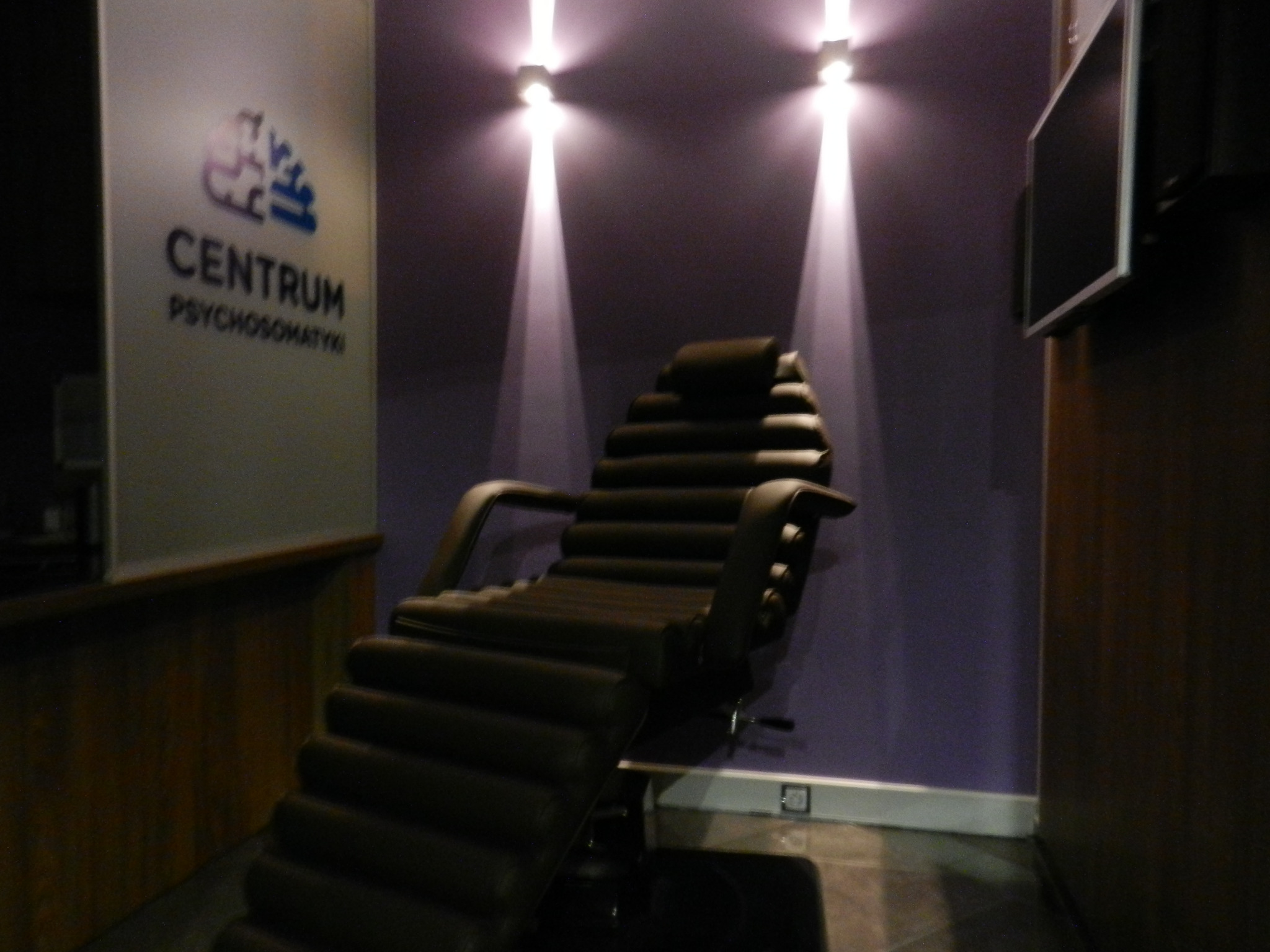 Wnętrze gabinetu z fotelem do terapii, oświetlonym punktowymi lampami na fioletowej ścianie, obok logo Centrum Psychosomatyki.