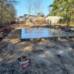 Świeżo wylany fundament płyty betonowej z widocznym szalunkiem, na piaszczystym terenie budowy z narzędziami w wiadrze w pierwszym planie i zabudową jednorodzinną w tle.