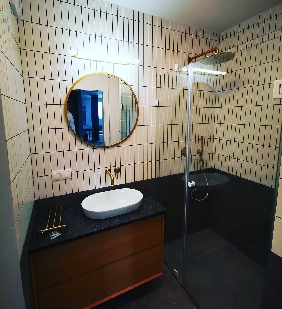 Nowoczesna łazienka z białymi płytkami ułożonymi pionowo z czarnymi fugami, okrągłe lustro w złotej ramie nad owalną umywalką nablatową, kabina prysznicowa ze szklaną ścianką i deszczownicą...