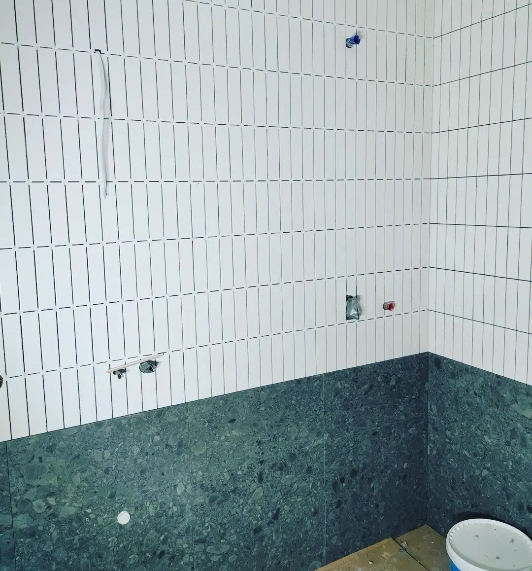 Fragment łazienki w trakcie wykańczania: białe, pionowe płytki ułożone na górnej części ścian, kontrastujące z ciemnoszarą, kamienną mozaiką w dolnej partii. Widoczne wyprowadzenia instalacji...