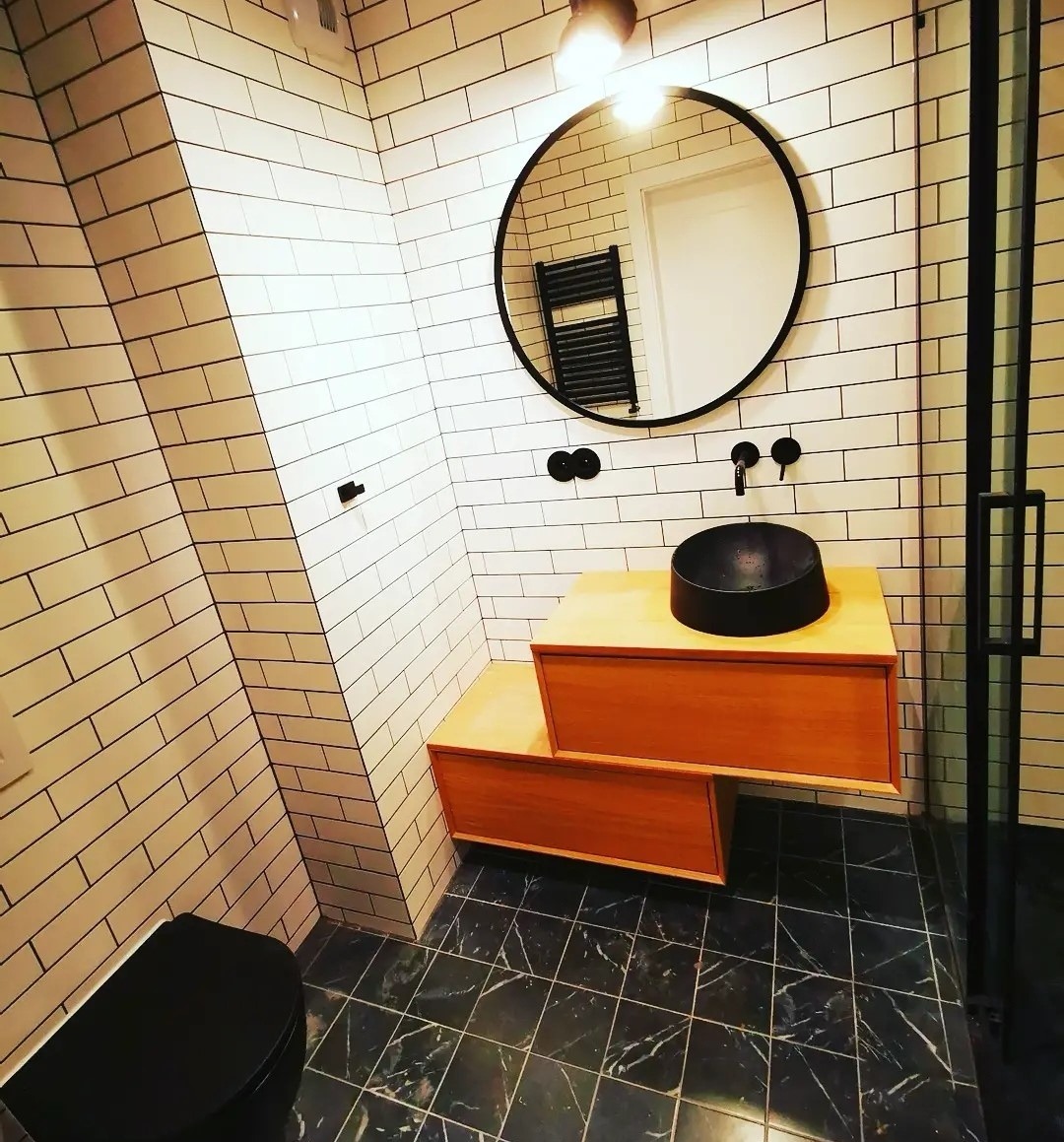 Nowoczesna łazienka z białą, skośną glazurą, czarną armaturą, okrągłym lustrem i drewnianą szafką pod umywalką, wykończona marmurową podłogą.