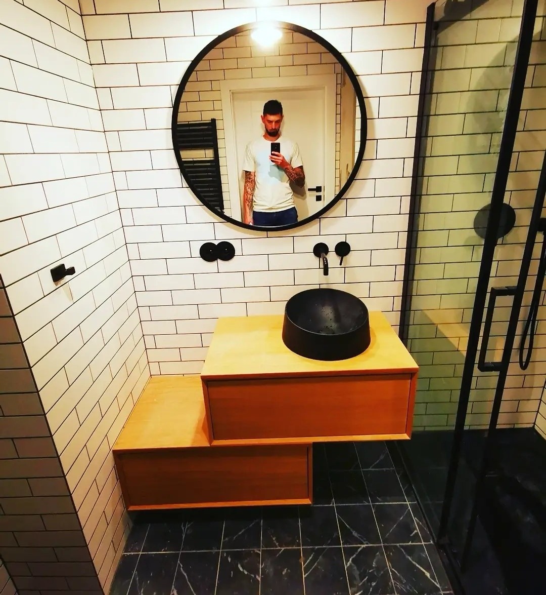 Nowoczesna łazienka z białymi, prostokątnymi płytkami, czarną umywalką nablatową na drewnianej szafce, czarną armaturą i okrągłym lustrem, w którym odbija się osoba robiąca zdjęcie.