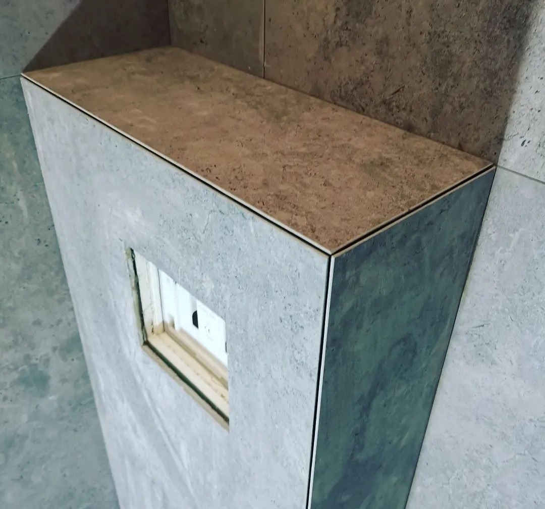 Półka w łazience wyłożona płytkami w odcieniach szarości i beżu, z widocznym otworem na instalację.