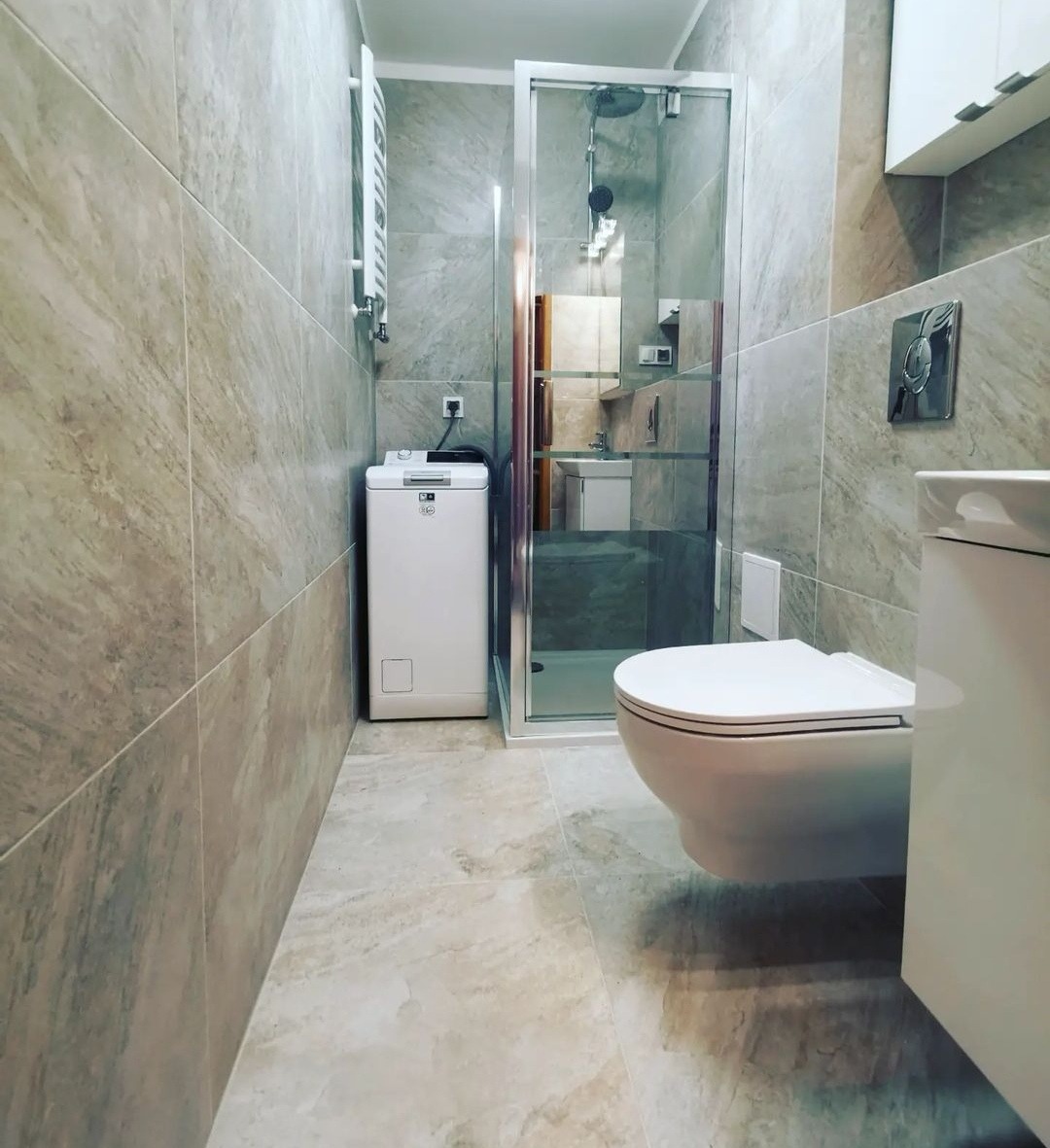 Wykończona łazienka z kabiną prysznicową, wiszącą toaletą, pralką i grzejnikiem drabinkowym, wyłożona płytkami imitującymi marmur w odcieniach szarości.