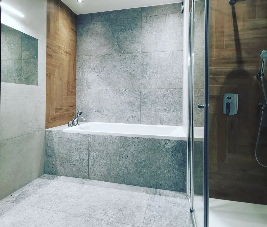 Nowoczesna łazienka z wanną obudowaną szarymi płytkami imitującymi beton, drewnopodobnymi panelami na ścianie i kabiną prysznicową z przezroczystego szkła.