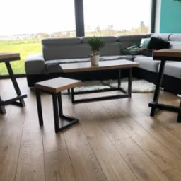 Nowoczesny salon z drewnianą podłogą, szarą sofą, stolikiem kawowym z metalową ramą i dwoma wysokimi stołkami barowymi z drewnianymi siedziskami i czarnymi, metalowymi nogami o nietypowym kształcie.