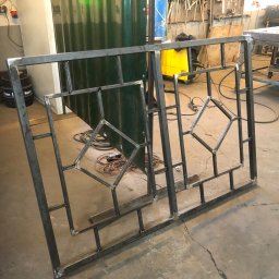 Piotr witkowski - Dwa surowe, spawane panele z metalowych profili o geometrycznym wzorze rombów i prostokątów, ustawione w warsztacie na betonowej posadzce, z widocznym sprzętem spawalniczym i narzędziami w tle.