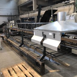 Piotr witkowski - Wnętrze hali produkcyjnej z konstrukcją stalową w trakcie budowy, widoczna drewniana paleta na pierwszym planie i elementy wentylacji pod sufitem.