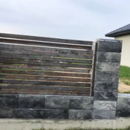 Montaż poziomej, metalowej bramy wjazdowej z elementami z szarej kostki betonowej, widoczny fragment domu z ciemną dachówką i pracownik w trakcie montażu.