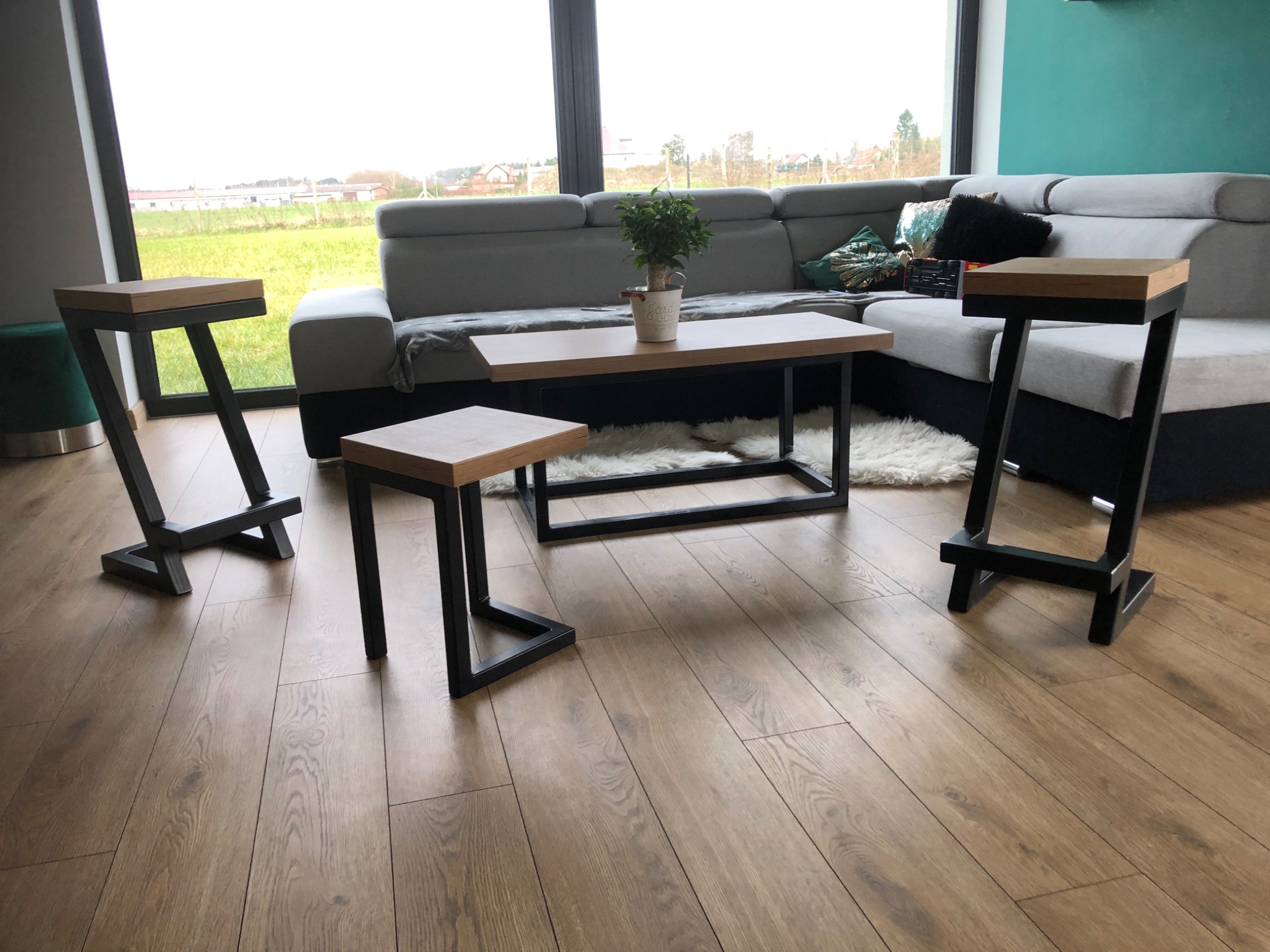 Nowoczesny salon z drewnianą podłogą, szarą sofą, stolikiem kawowym z metalową ramą i dwoma wysokimi stołkami barowymi z drewnianymi siedziskami i czarnymi, metalowymi nogami o nietypowym kształcie.