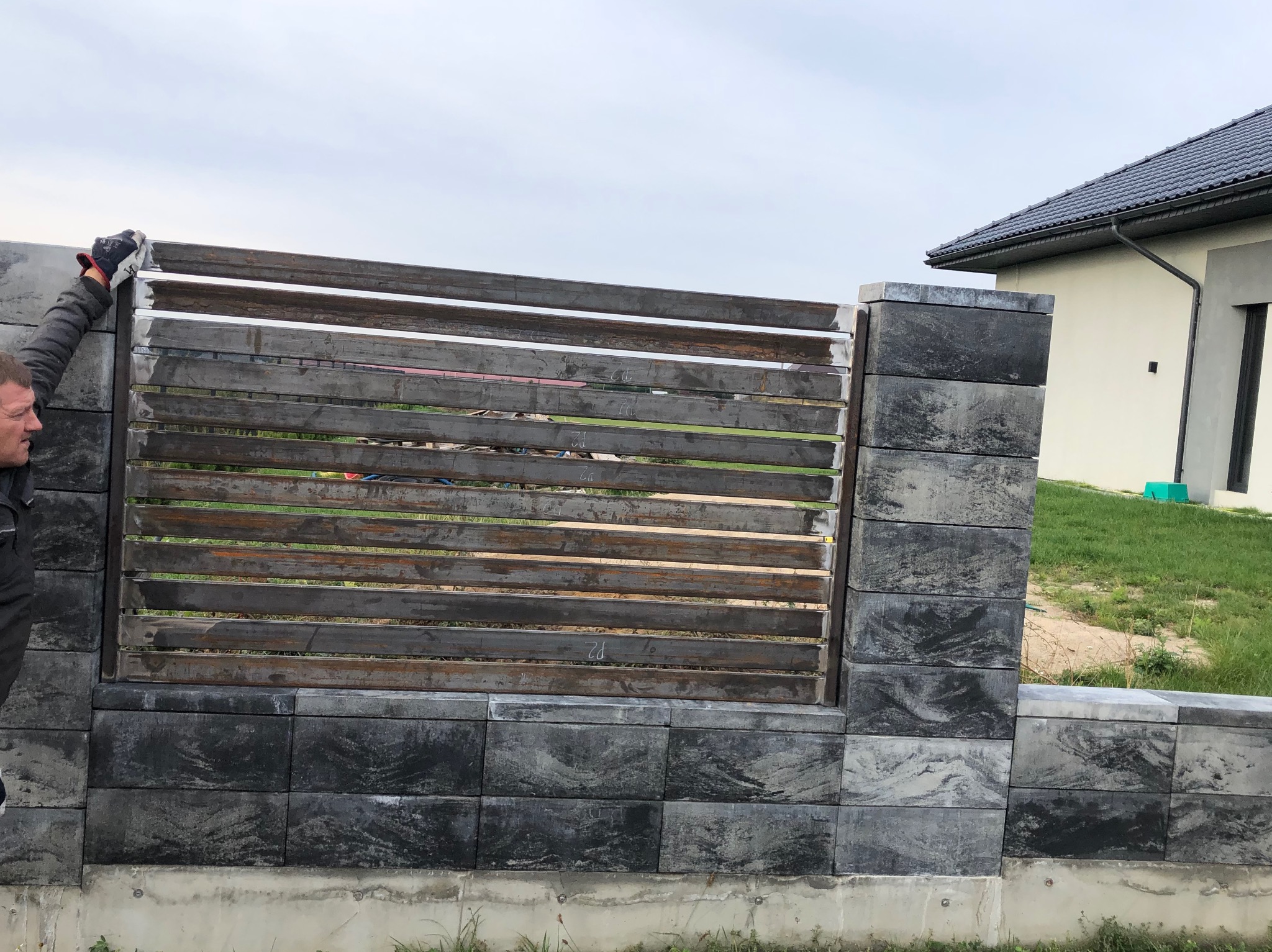 Montaż poziomej, metalowej bramy wjazdowej z elementami z szarej kostki betonowej, widoczny fragment domu z ciemną dachówką i pracownik w trakcie montażu.