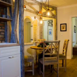 Jasne wnętrze mieszkania z elementami rustykalnymi: drewniane belki konstrukcyjne, ceglana ściana z półkami i okrągły stół z krzesłami w stylu vintage.