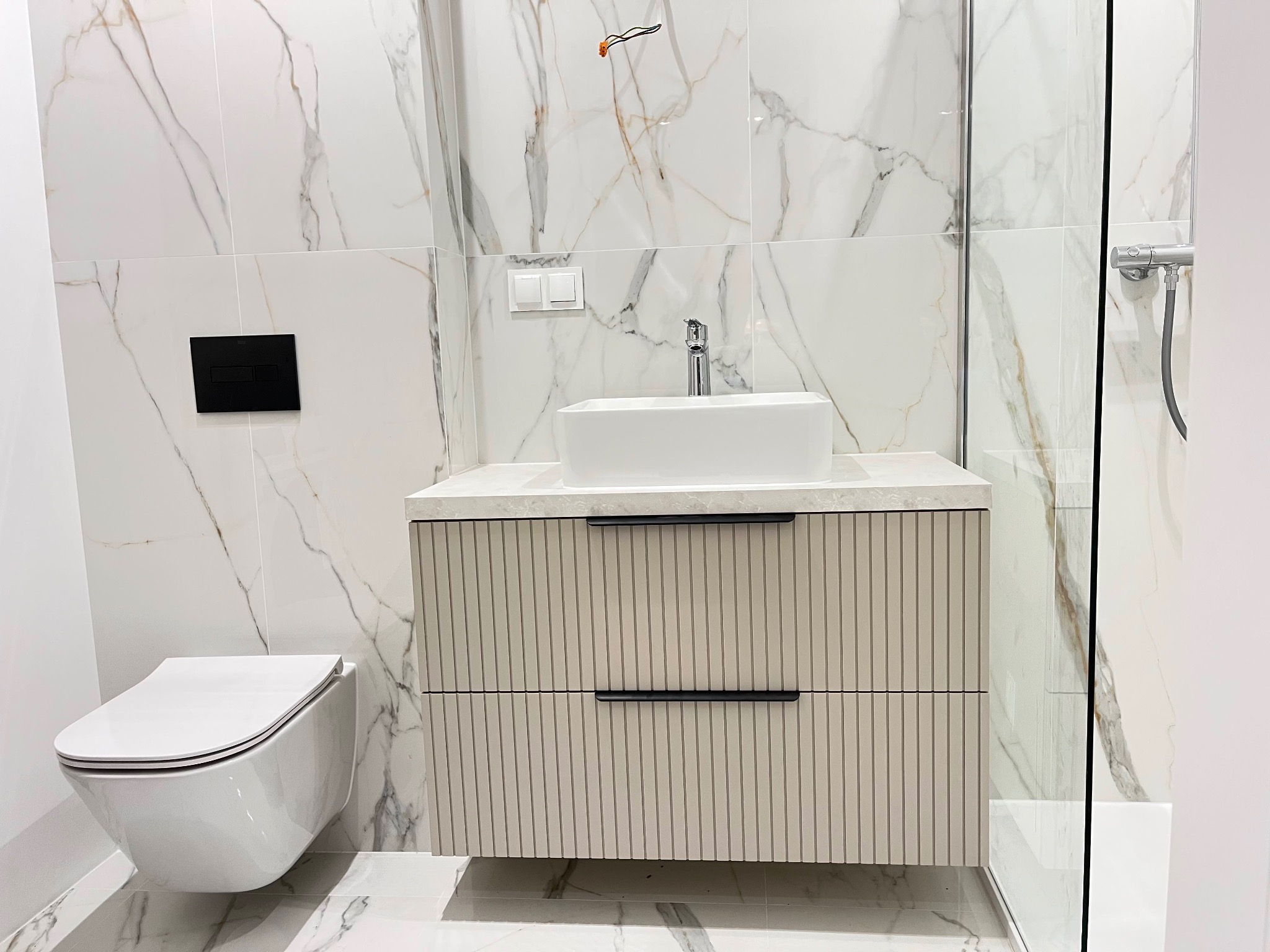 Nowoczesna łazienka z marmurowymi płytkami, wiszącą toaletą i szafką pod umywalkę z ryflowanymi frontami. Minimalistyczny design, chromowana armatura.