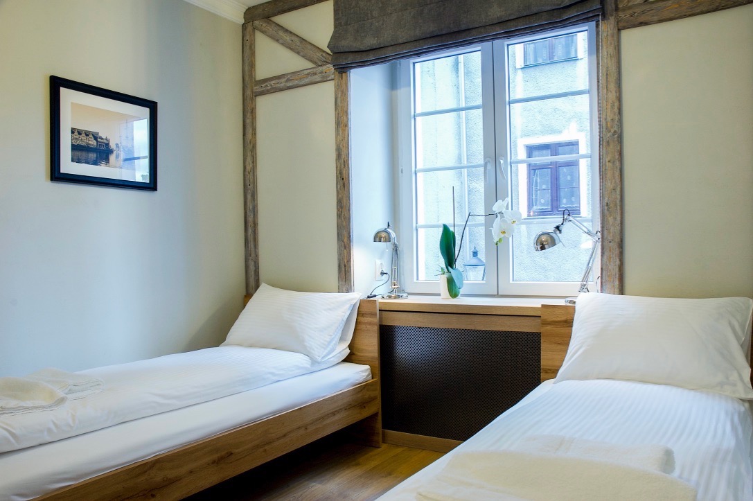 Jasny pokój gościnny z dwoma pojedynczymi łóżkami, drewnianą ramą okienną i dekoracyjną drewnianą konstrukcją na ścianie, lampkami nocnymi i białymi orchideami na parapecie.