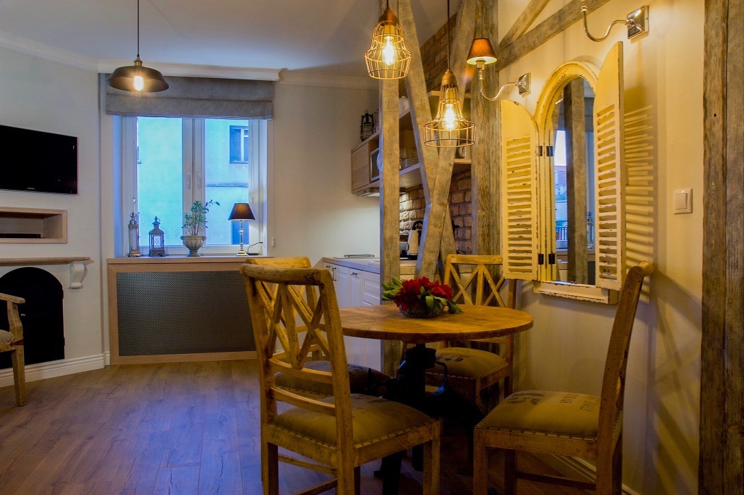 Jasne wnętrze jadalni z okrągłym drewnianym stołem, krzesłami z krzyżowym oparciem i dekoracyjnymi lampami wiszącymi w stylu industrialnym, uzupełnione rustykalnymi elementami architektonicznymi...