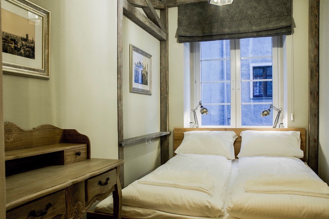 Jasny pokój w Gdańsku z dwoma łóżkami pojedynczymi, drewnianą ramą, biurkiem z rzeźbieniami i oknem z widokiem na budynek.