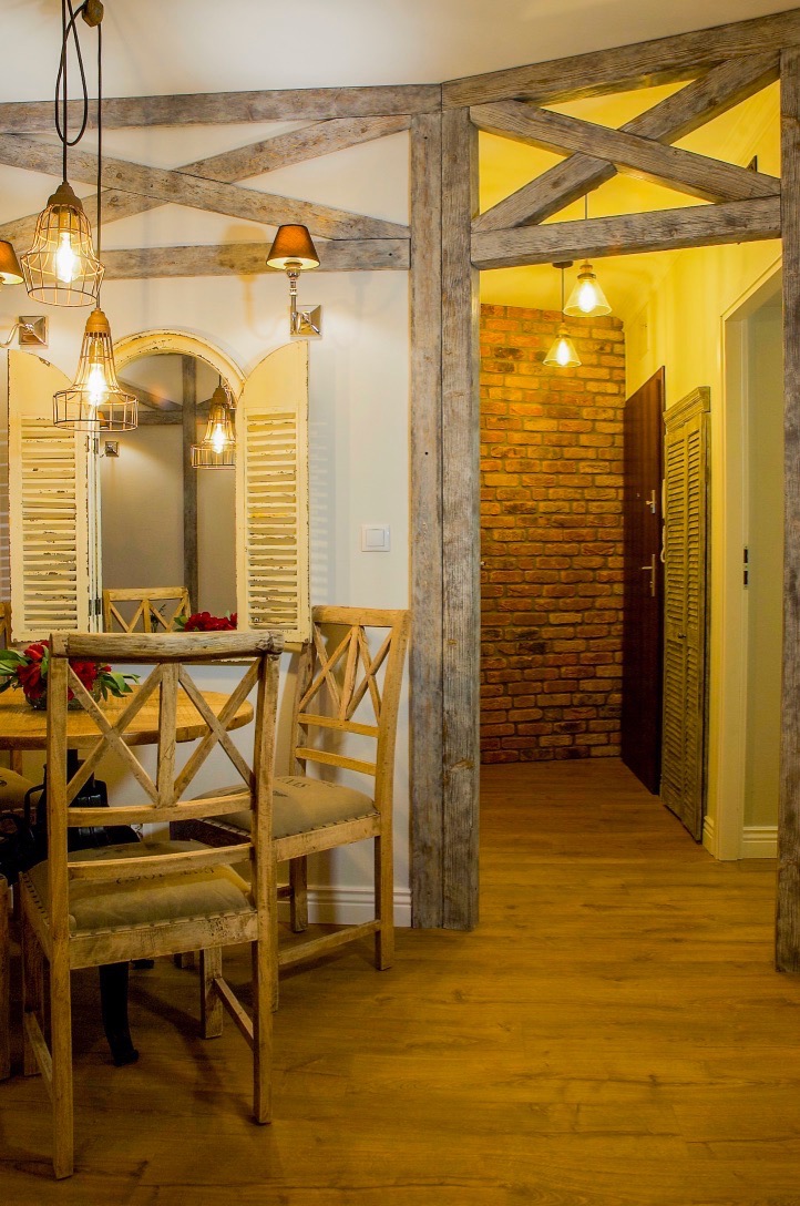 Rustykalne wykończenie wnętrza: drewniane belki, ceglana ściana, lustro z okiennicami i krzesła z surowego drewna, oświetlenie w stylu industrialnym.