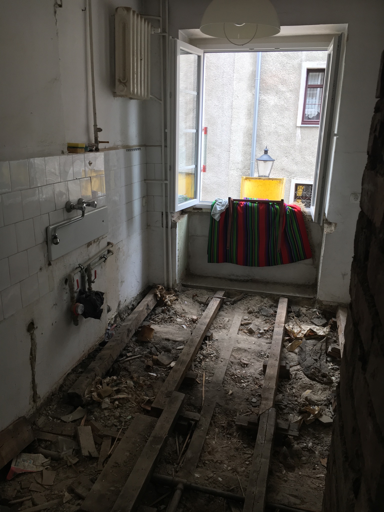 Wnętrze łazienki w trakcie demontażu, widoczne odsłonięte deski podłogowe, stare płytki na ścianach, instalacja wodna i okno z widokiem na budynek sąsiedni.