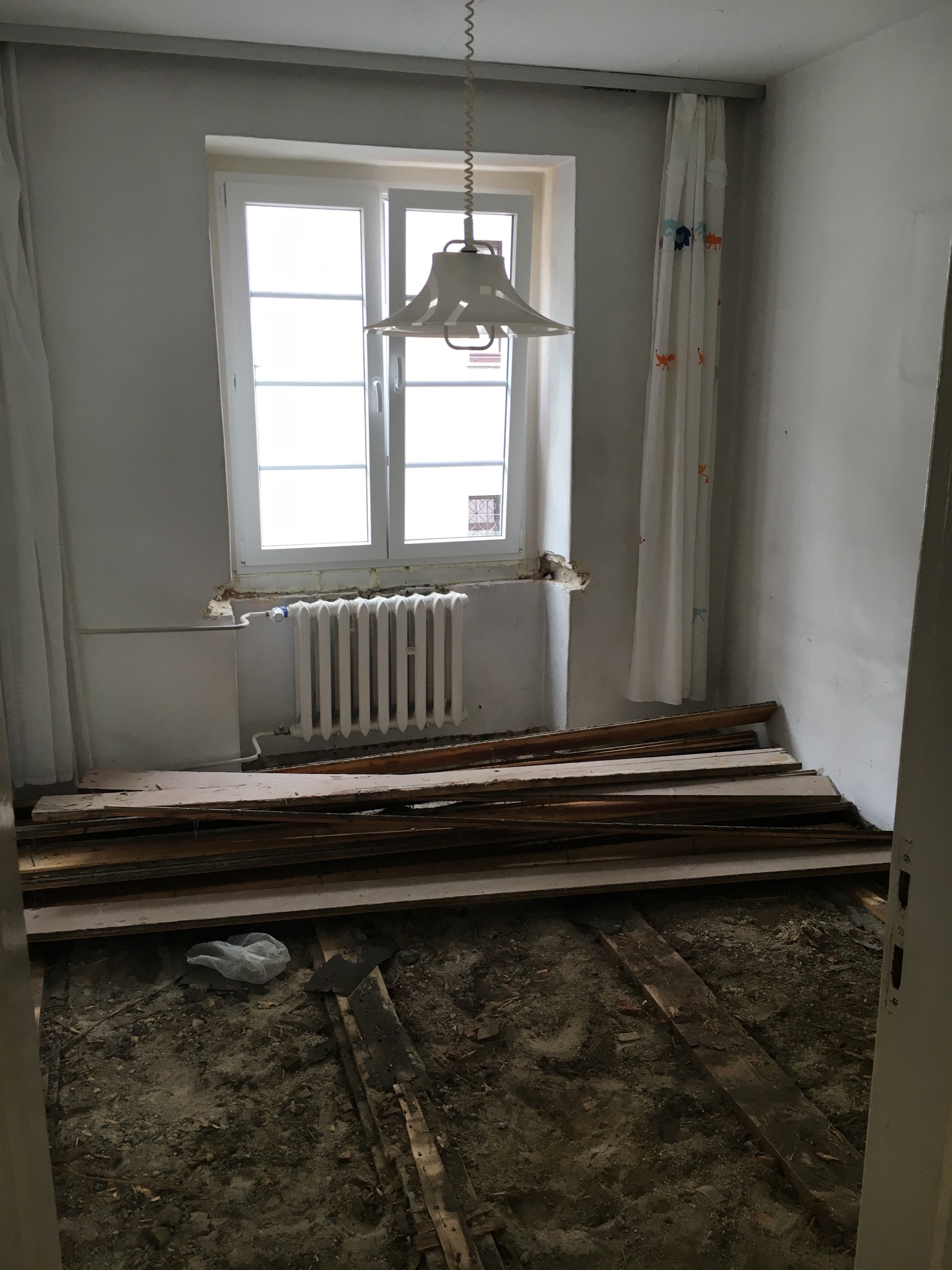 Pokój w trakcie demontażu podłogi, widoczne stare deski, gruz i odsłonięte legary, białe ściany, okno z grzejnikiem, zasłony z motywami zwierzęcymi.