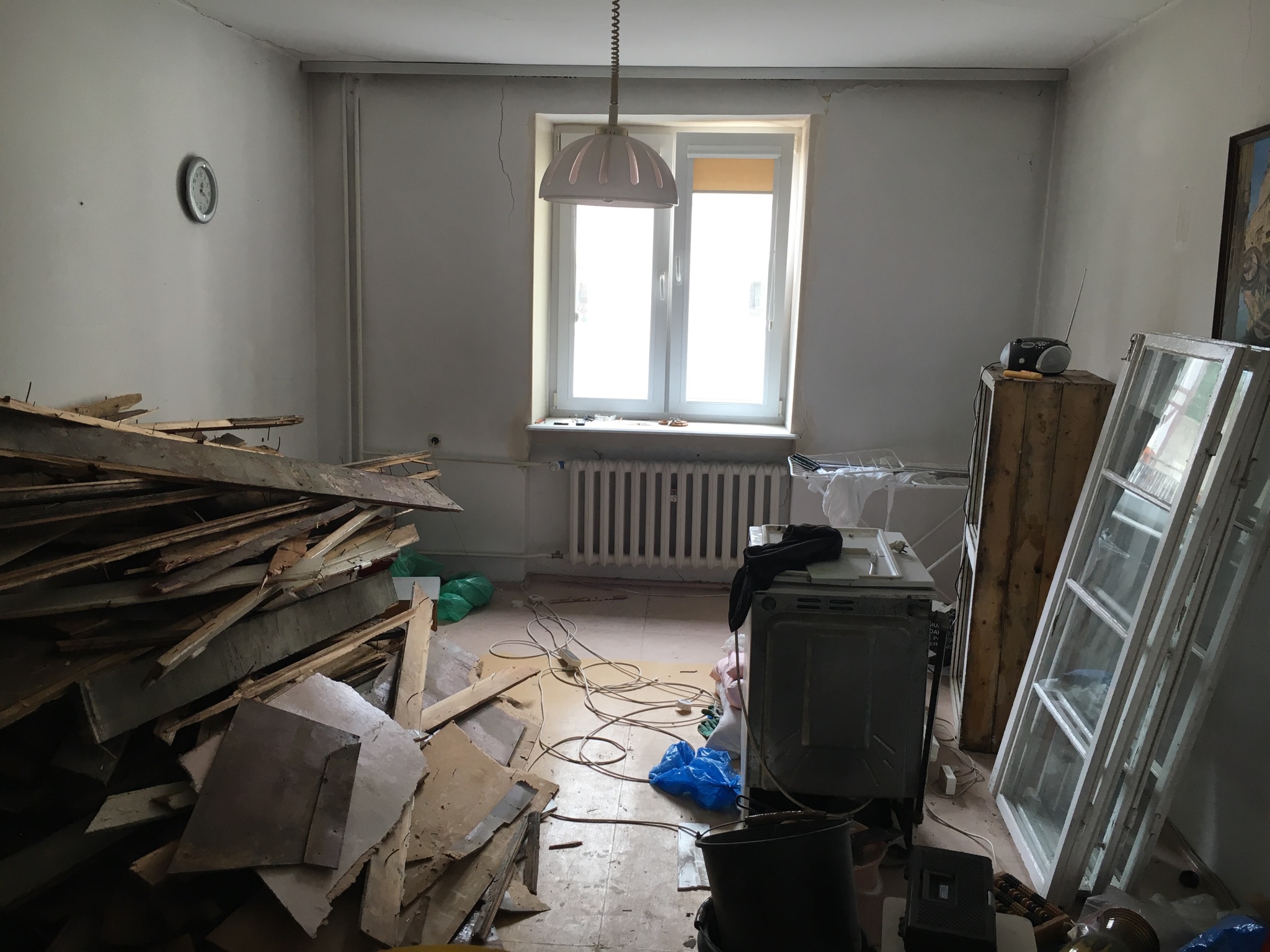 Pokój w trakcie demontażu: sterta drewnianych elementów, odłączony sprzęt AGD, stare okna oparte o ścianę, widoczny bałagan i resztki materiałów.