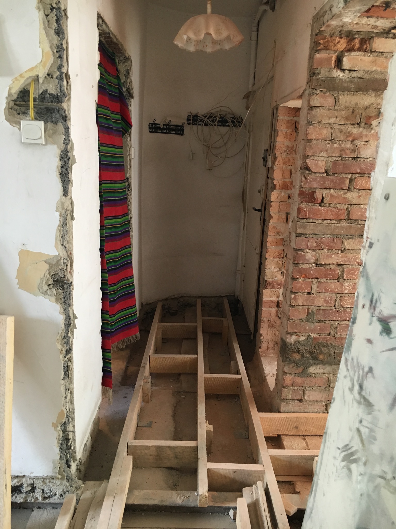 Wnętrze w trakcie remontu: odsłonięta cegła, fragmenty drewnianej konstrukcji podłogi, ściany z usuniętym tynkiem, wiszący kabel elektryczny i kolorowa tkanina w przejściu.