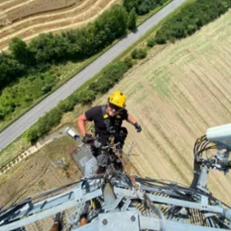 Pracownik w uprzęży i żółtym kasku na szczycie metalowej konstrukcji wieży telekomunikacyjnej, widok z góry na pola i drogę, słoneczny dzień.