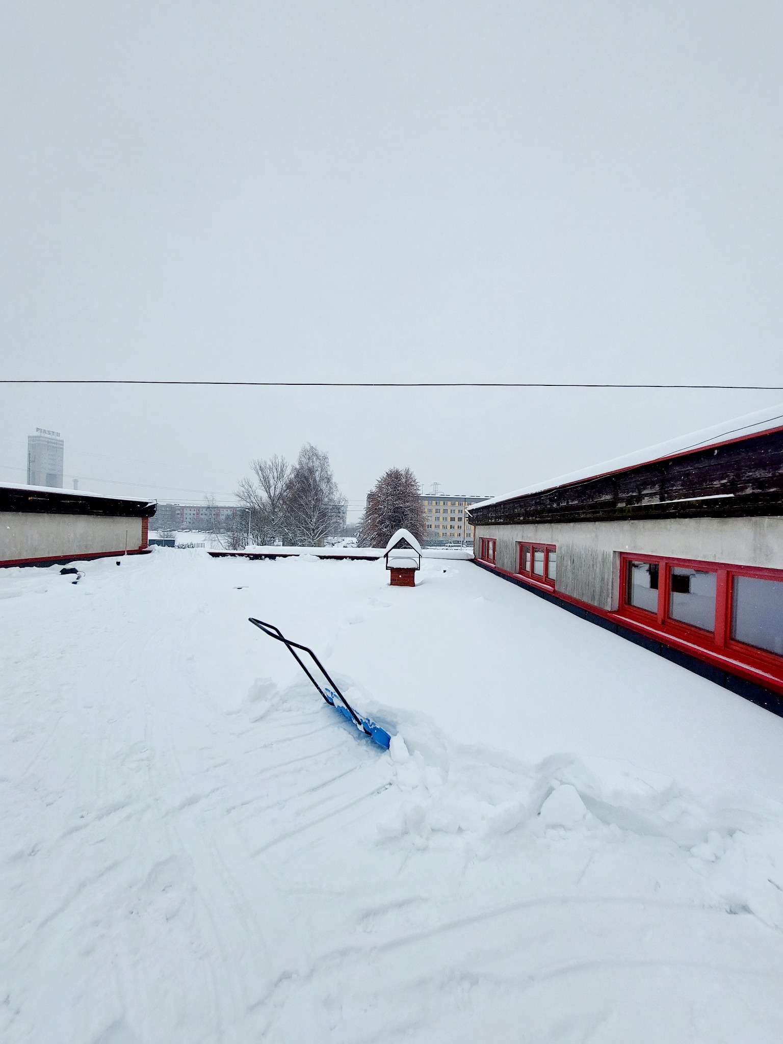 Śnieżny dach budynku z widocznymi śladami odśnieżania i niebieską łopatą do śniegu, w tle zabudowa miejska pod szarym niebem.