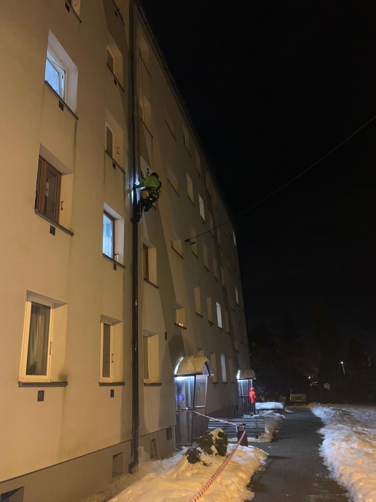 Pracownik na linach zabezpieczających, w jaskrawej kamizelce, czyści rynnę na elewacji bloku mieszkalnego nocą, podświetlając sobie latarką czołową; w tle ośnieżony chodnik i budynek.