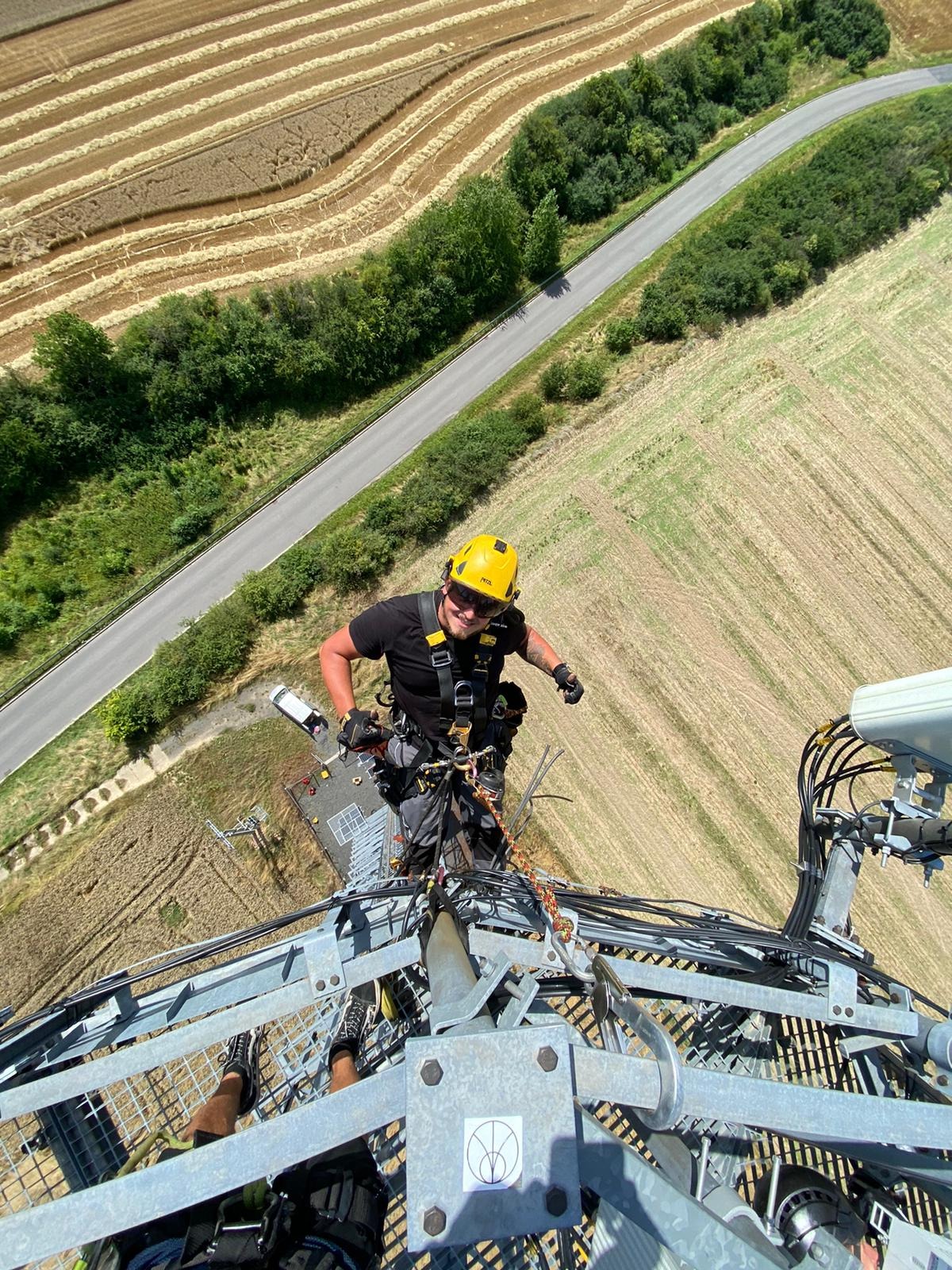 Pracownik w uprzęży i żółtym kasku na szczycie metalowej konstrukcji wieży telekomunikacyjnej, widok z góry na pola i drogę, słoneczny dzień.