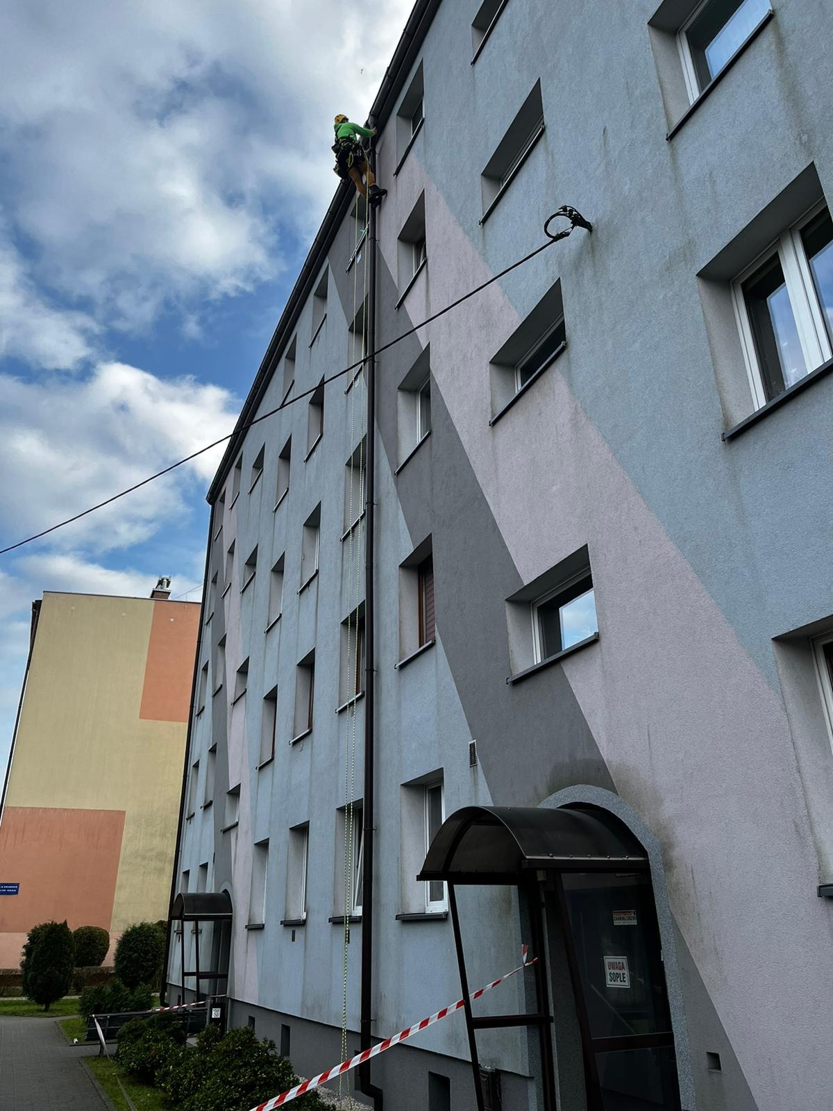 Pracownik w uprzęży linowej przy elewacji budynku wielopiętrowego z geometrycznym wzorem, zabezpieczony taśmą ostrzegawczą przy wejściu.