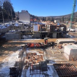 OstBudHousDom - Widok z góry na plac budowy w zimowy dzień: zbrojenie, szalunki, betonowe fundamenty i elementy konstrukcyjne w trakcie prac, z widocznym sprzętem budowlanym i częściowo ośnieżonym terenem.