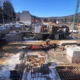 Widok z góry na plac budowy w zimowy dzień: zbrojenie, szalunki, betonowe fundamenty i elementy konstrukcyjne w trakcie prac, z widocznym sprzętem budowlanym i częściowo ośnieżonym terenem.