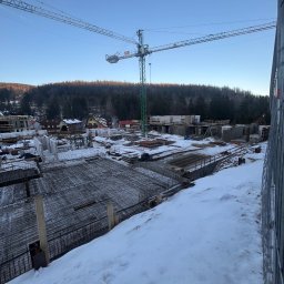 OstBudHousDom - Zimowy krajobraz placu budowy z widoczną konstrukcją zbrojeniową fundamentów, żurawiem budowlanym w tle i ośnieżonym terenem.