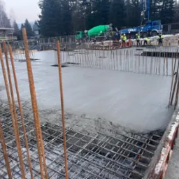 Wylewanie betonu na zbrojone fundamenty z widocznymi pracownikami w odblaskowych kamizelkach, pompą do betonu CIFA i betoniarkami w tle.