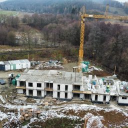 OstBudHousDom - Budowa dwukondygnacyjnego budynku z białego bloczka, widok z góry. Na dachu widoczne zbrojenia, w tle żuraw budowlany i zalesione wzgórza. Częściowo ośnieżony teren.