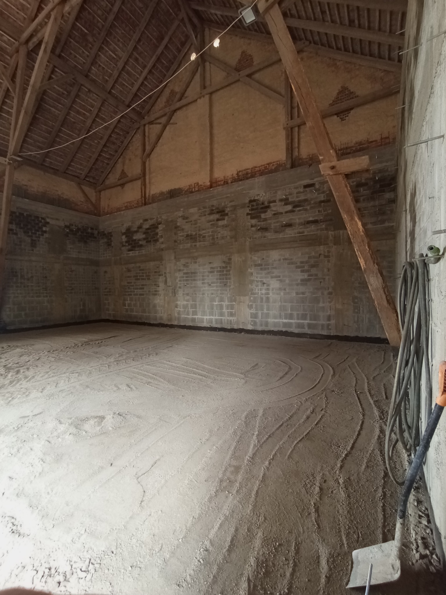Wnętrze budynku w trakcie prac: przygotowane podłoże pod wylewkę, widoczne ślady po rozprowadzaniu masy, ceglane ściany i drewniana konstrukcja dachu, w rogu oparta szpadel i zwinięty wąż.