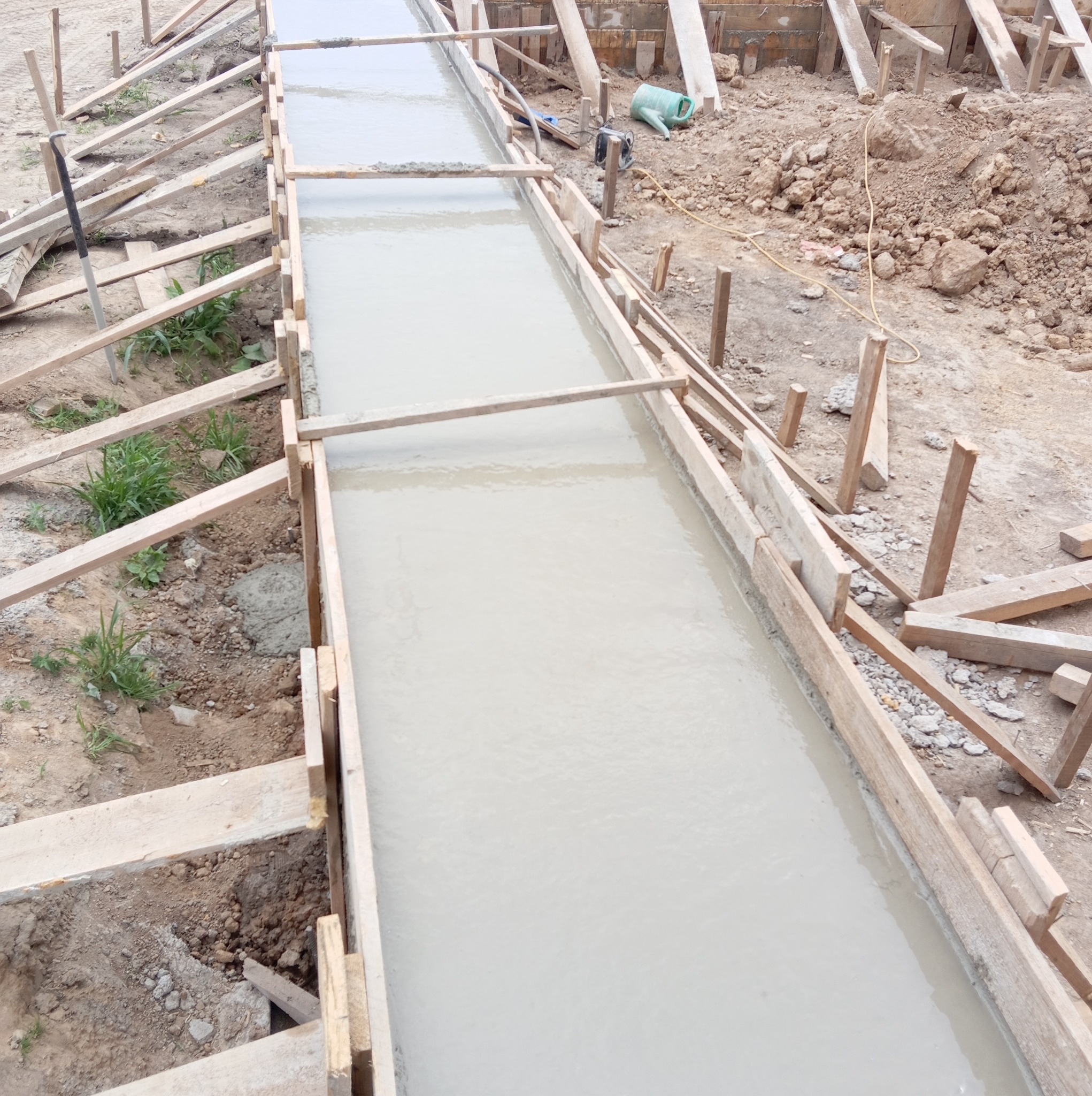 Wypełniony betonem wąski wykop z drewnianym szalunkiem, widoczne narzędzia i ziemia wokół.