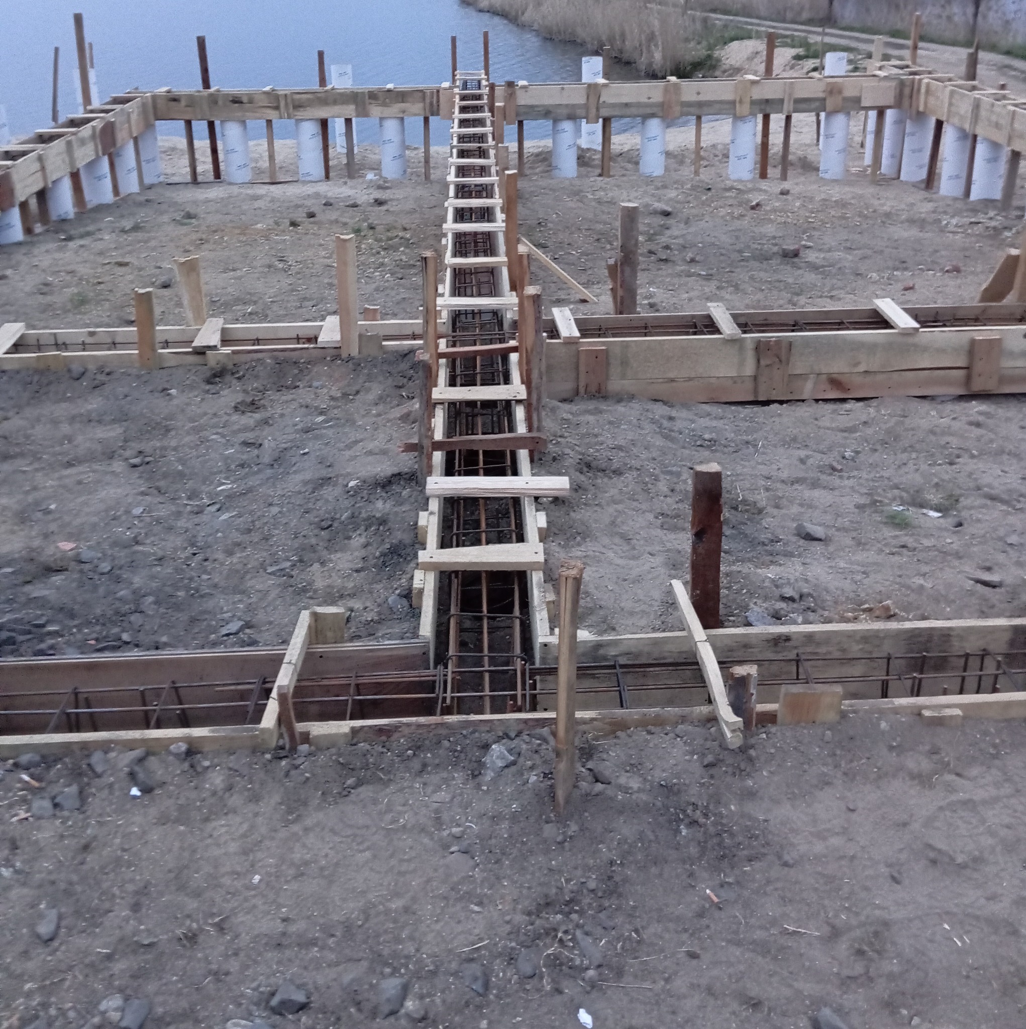 Przygotowanie terenu pod budowę: widoczne deskowanie i zbrojenie fundamentów, z rurami szalunkowymi w tle, na tle zbiornika wodnego.