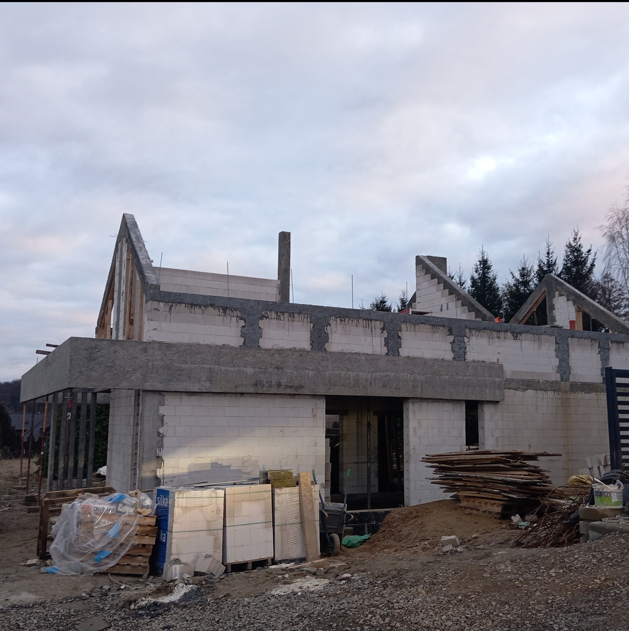 Surowy stan domu w trakcie budowy: ściany z bloczków, częściowo zalany strop, widoczne zbrojenia i drewniane elementy szalunków, materiały budowlane składowane obok.