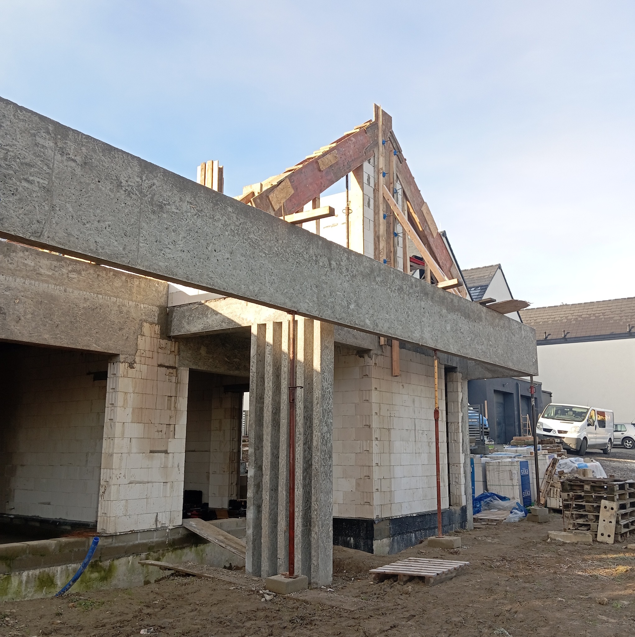 Wznoszenie ścian domu z bloczków silikatowych, widoczne elementy szalunku stropu i konstrukcja dachu w trakcie budowy na tle zabudowy jednorodzinnej.