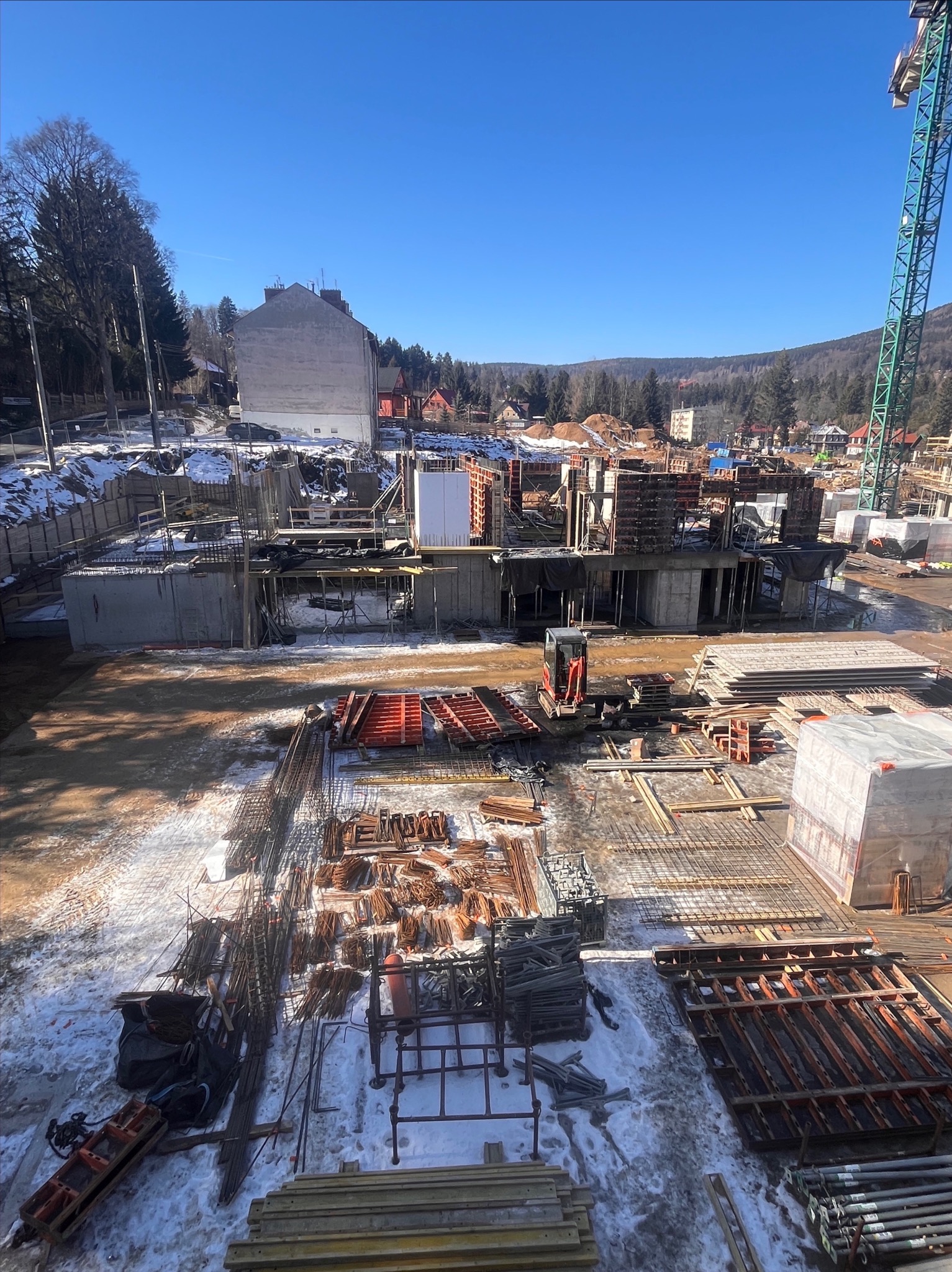 Widok z góry na plac budowy w zimowy dzień: zbrojenie, szalunki, betonowe fundamenty i elementy konstrukcyjne w trakcie prac, z widocznym sprzętem budowlanym i częściowo ośnieżonym terenem.