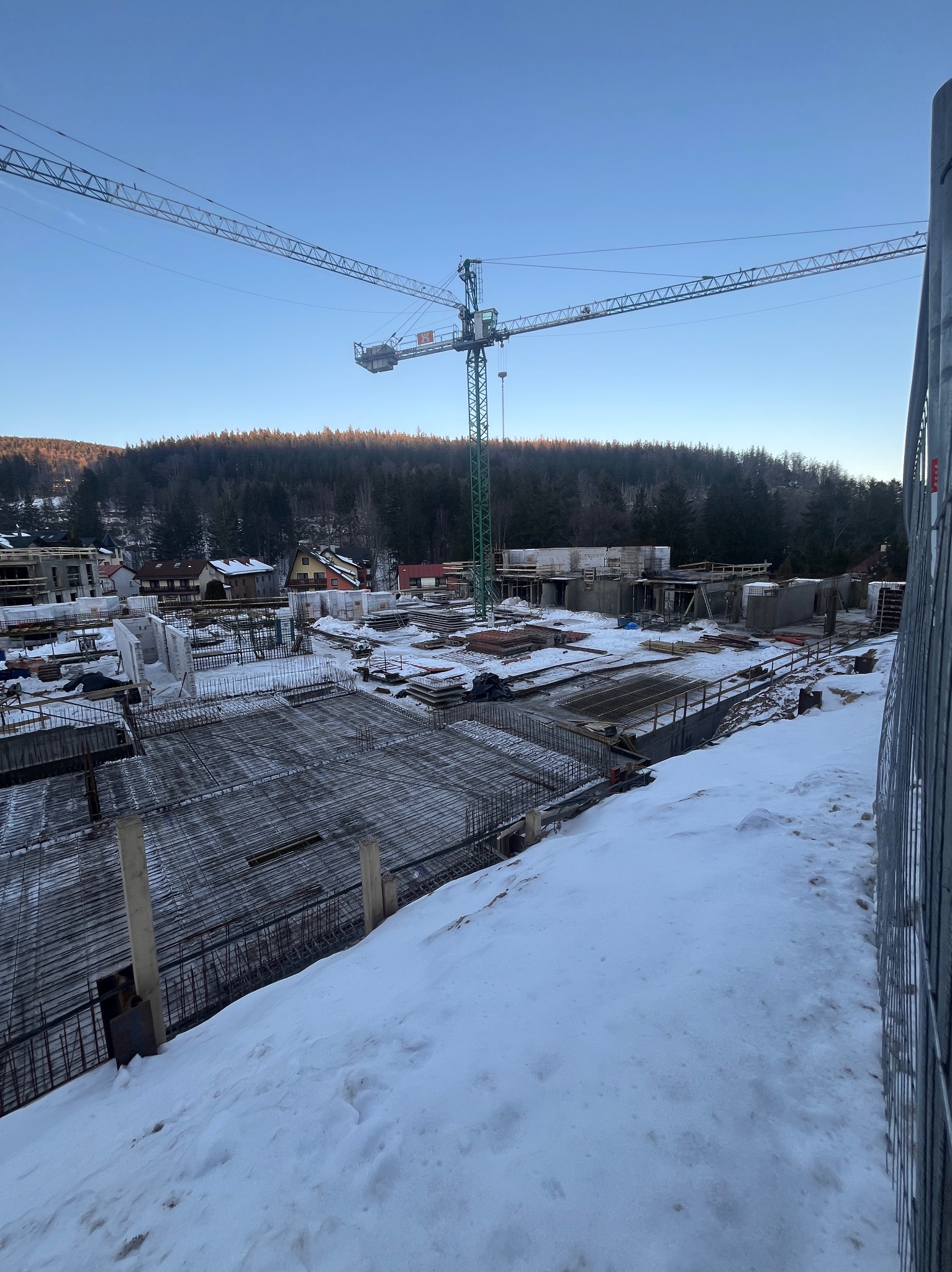 Zimowy krajobraz placu budowy z widoczną konstrukcją zbrojeniową fundamentów, żurawiem budowlanym w tle i ośnieżonym terenem.