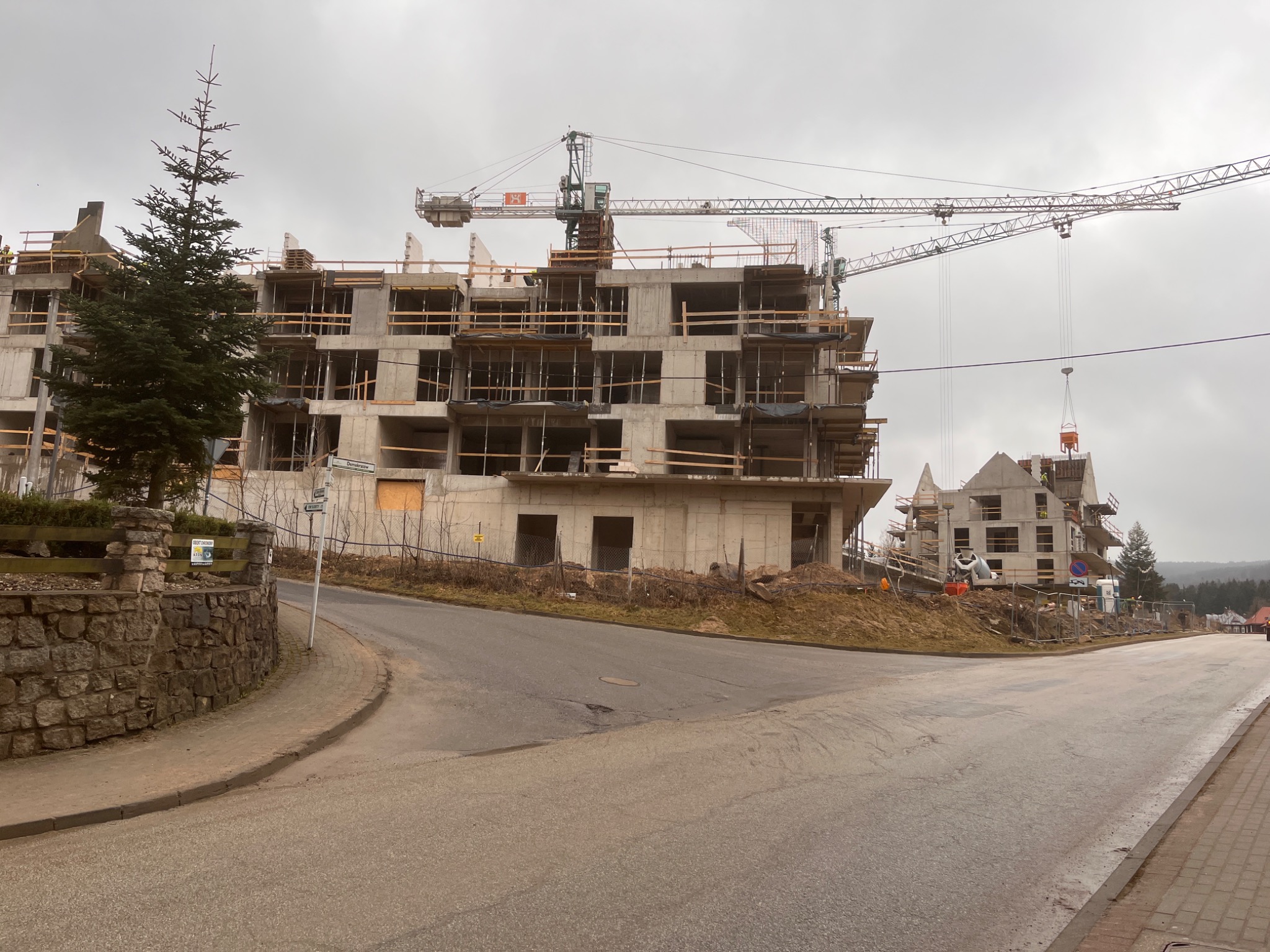 Wielopiętrowy budynek w trakcie wznoszenia z betonu, z widocznym szalunkiem i żurawiami budowlanymi na tle pochmurnego nieba. Na pierwszym planie skrzyżowanie dróg.