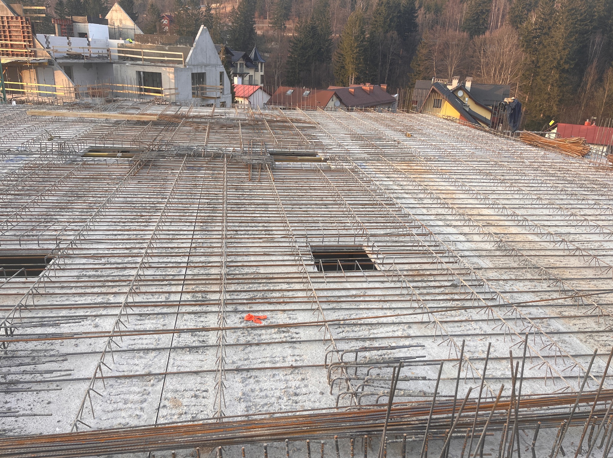 Zbrojenie płyty fundamentowej budynku w trakcie budowy, widok z góry na siatkę z prętów stalowych, z betonem w tle i fragmentami drewnianego szalunku oraz zabudową mieszkalną w oddali.