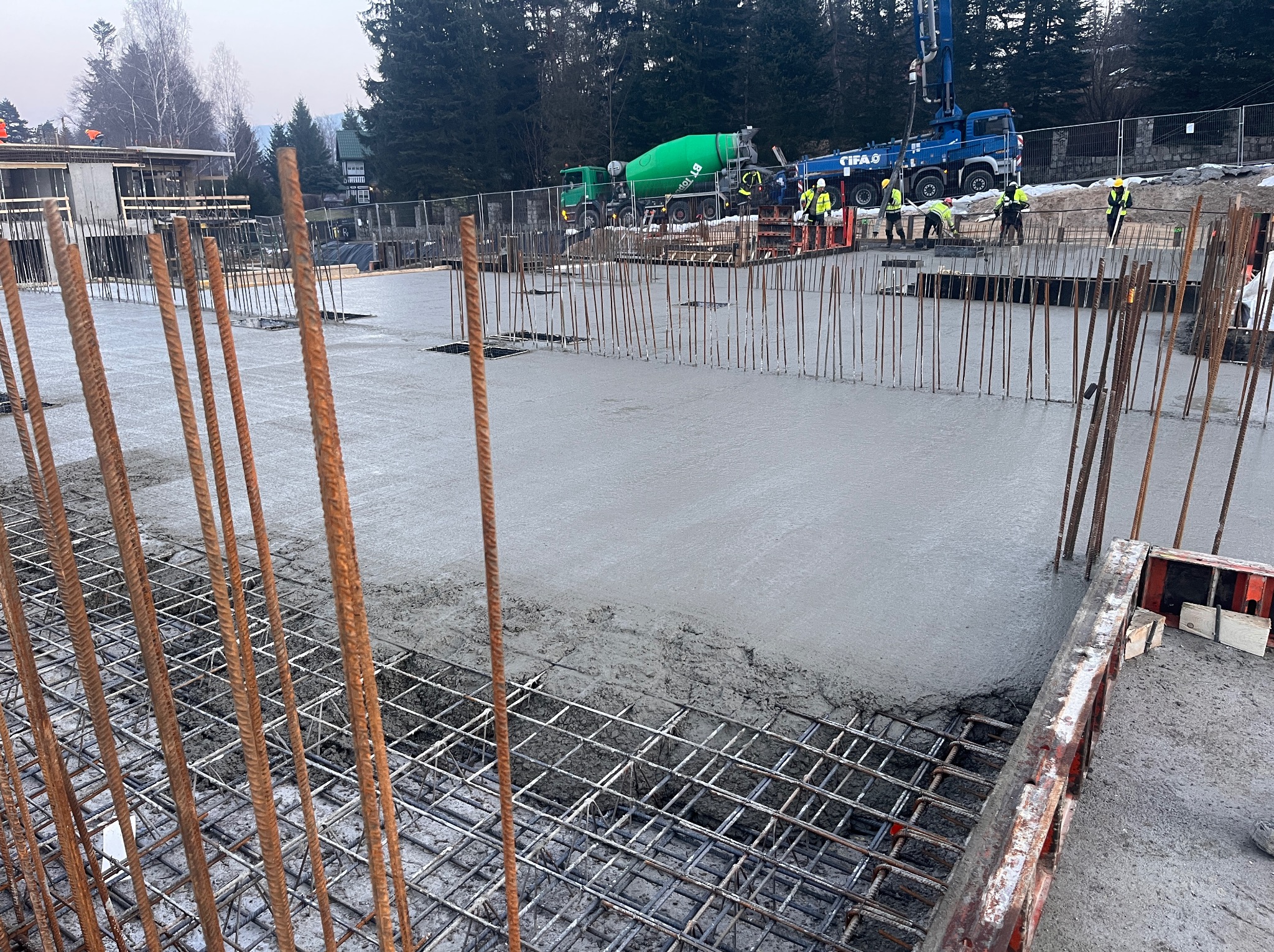 Wylewanie betonu na zbrojone fundamenty z widocznymi pracownikami w odblaskowych kamizelkach, pompą do betonu CIFA i betoniarkami w tle.