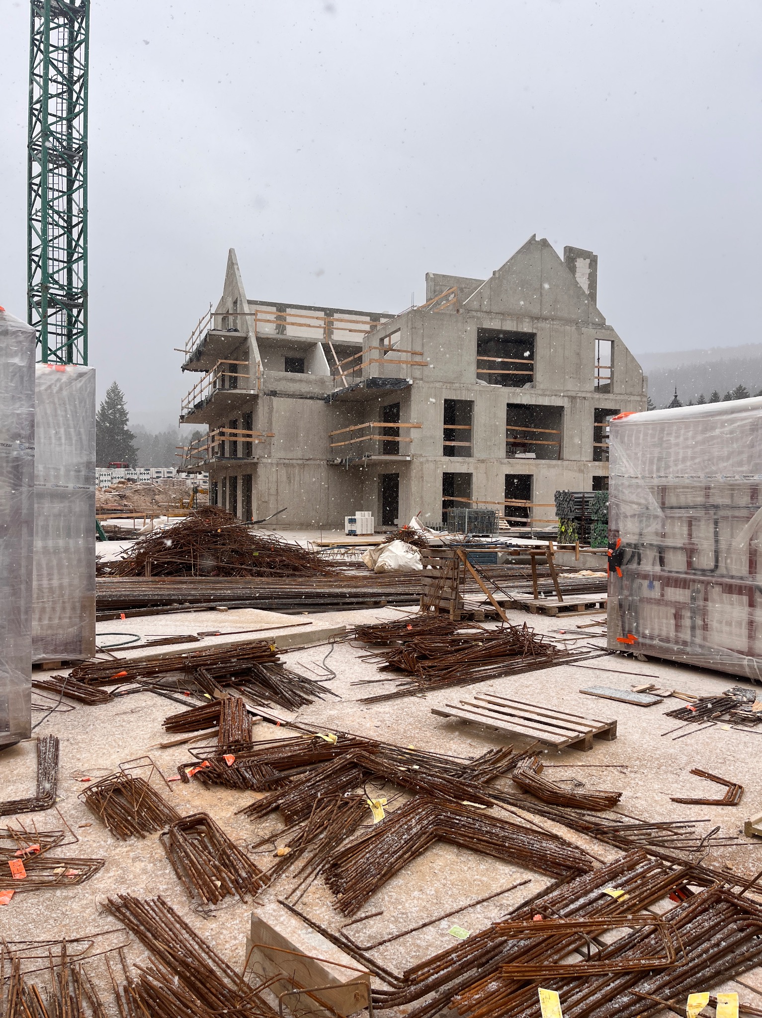 Surowy, trzypiętrowy budynek z betonu w trakcie budowy, widziany podczas opadów śniegu. Na pierwszym planie porozrzucane pręty zbrojeniowe z pomarańczowymi etykietami.