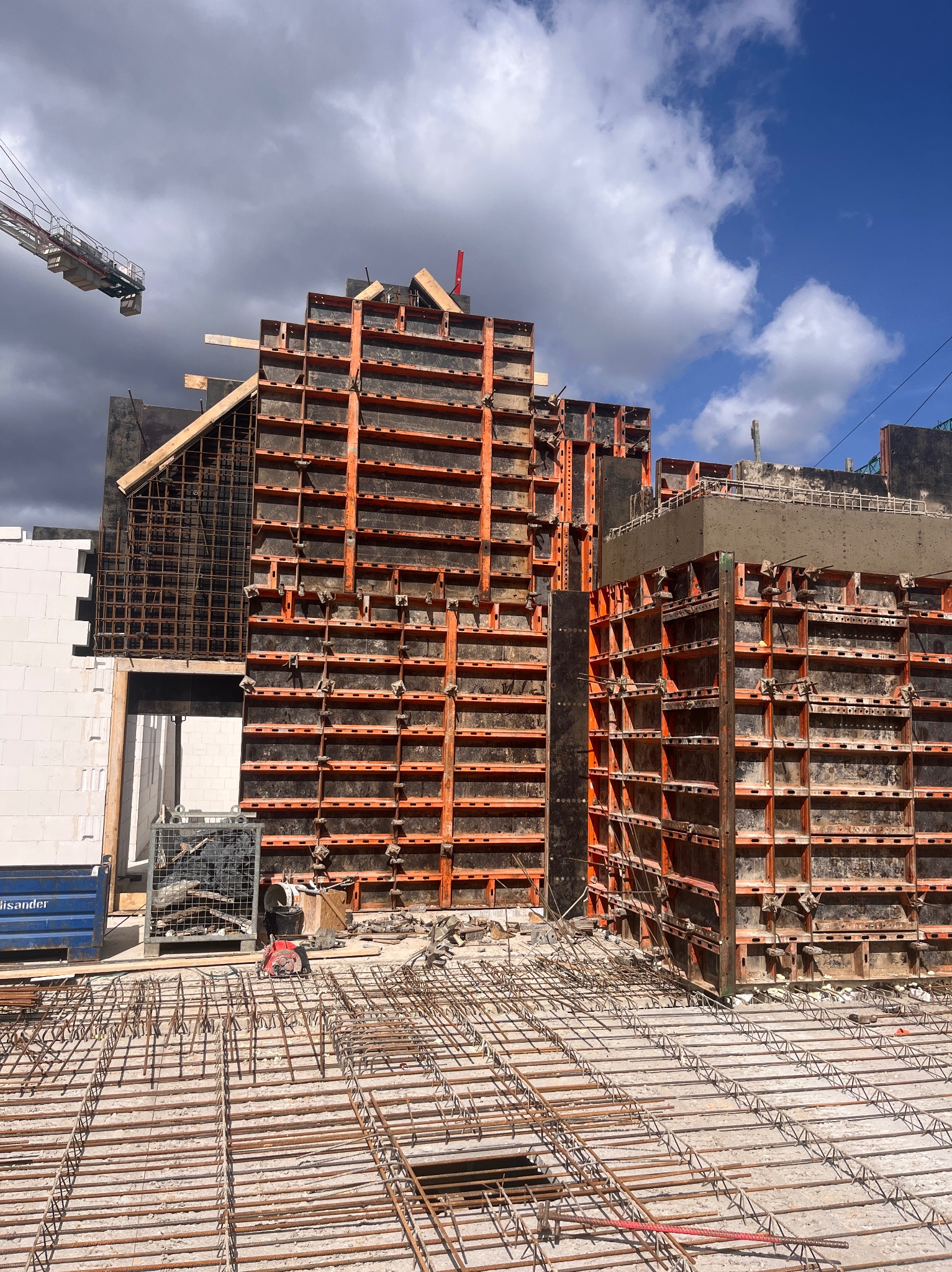 Szare szalunki metalowe z pomarańczowymi elementami konstrukcyjnymi, przygotowane pod wylanie betonu, widoczne zbrojenia i częściowo wymurowana ściana z białych bloczków oraz żuraw budowlany w tle.