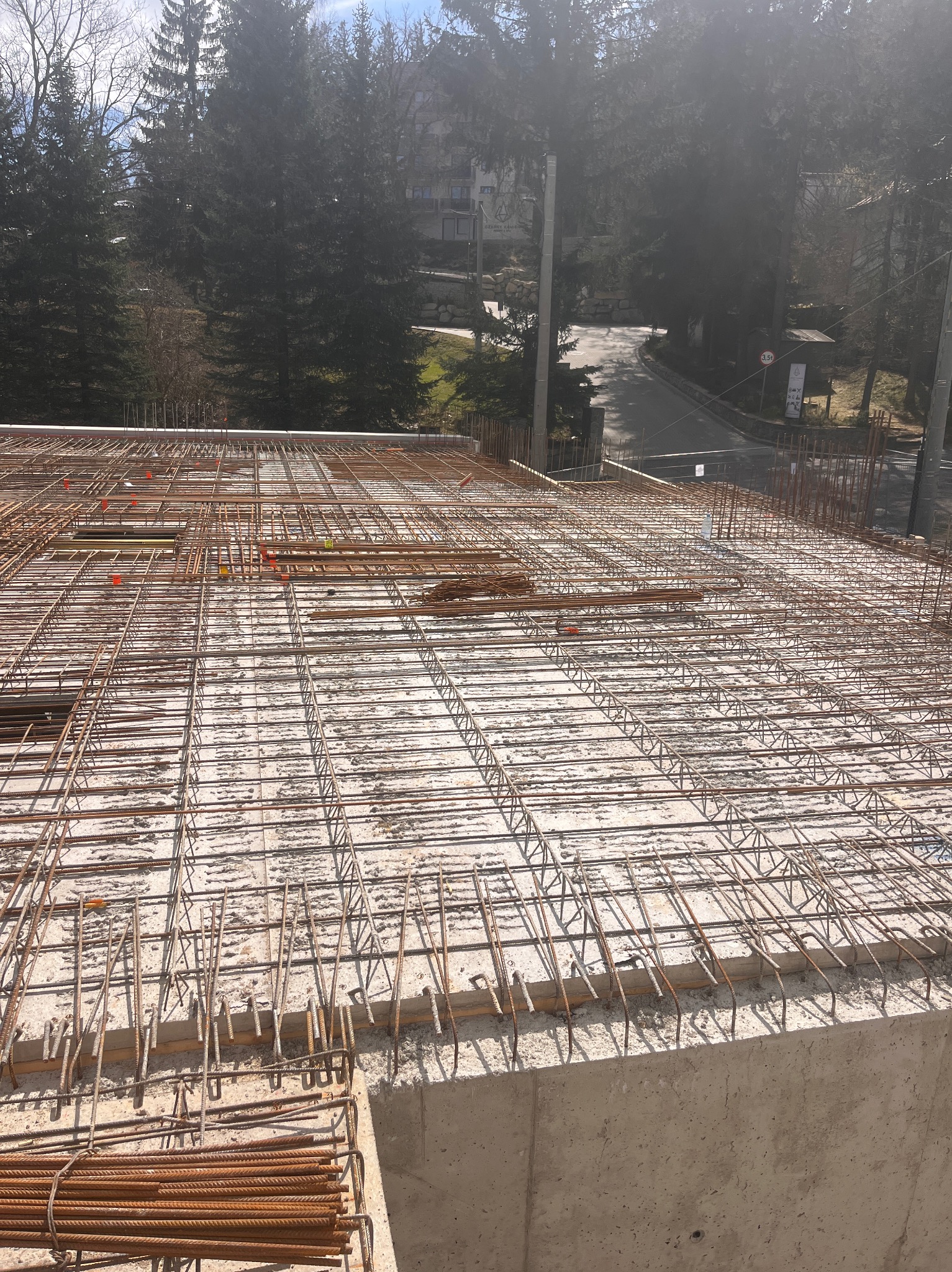 Zbrojenie stropu budynku w trakcie budowy, widok z góry na siatkę z prętów zbrojeniowych na betonowej konstrukcji, w tle drzewa i budynki.